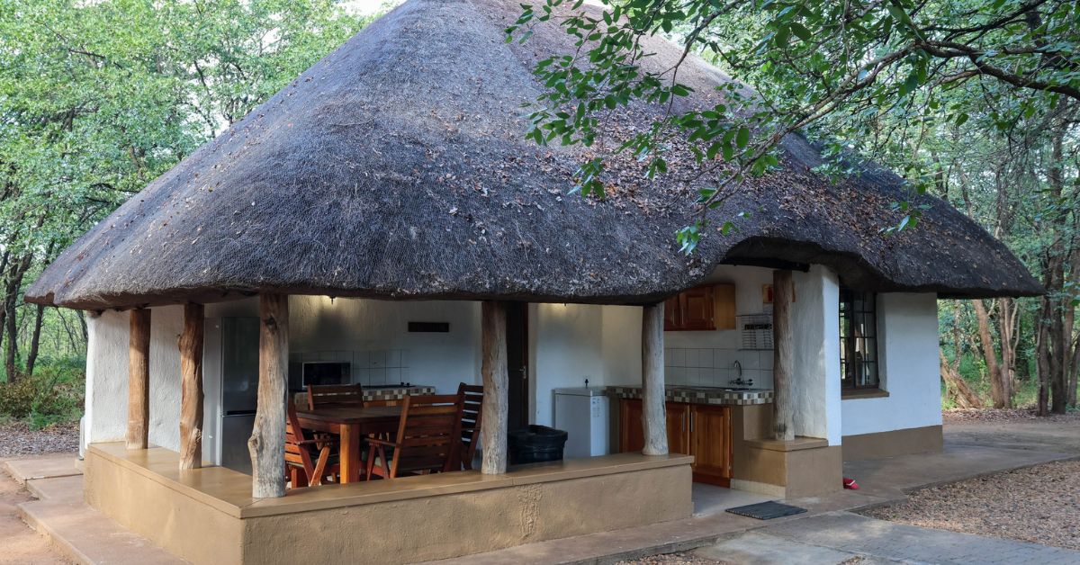 A white-walled safari chalet with a thick thatched roof and an open-air kitchen and dining area on the porch, surrounded by lush trees at Bateleur Bushveld Camp.