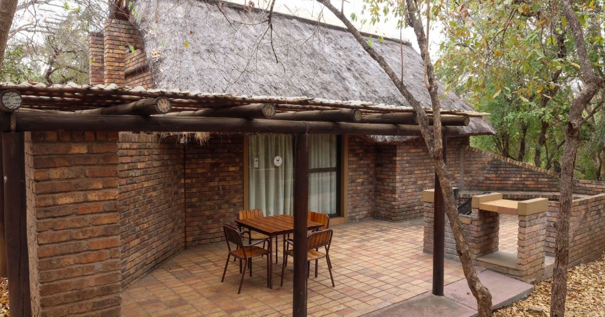 A face-brick safari chalet with a thatched roof and a paved patio area including a wooden table, chairs, and a private braai facility at Berg-en-Dal Rest Camp.