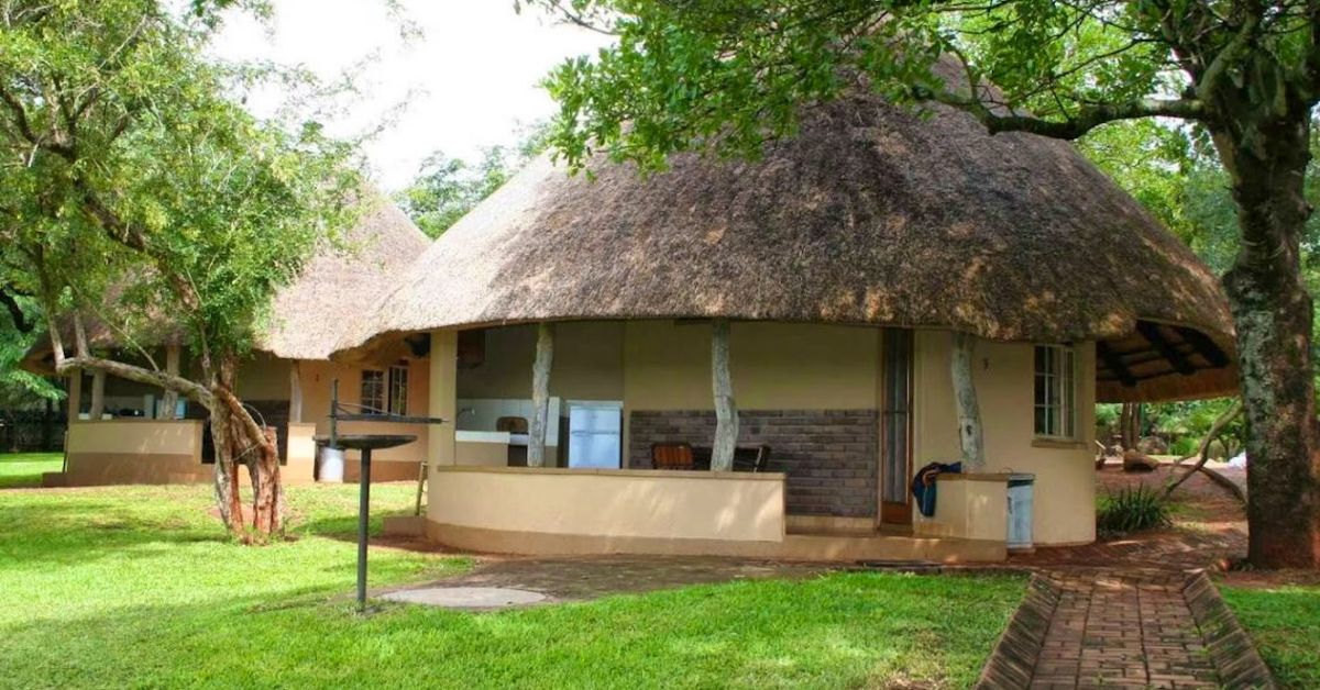 Two traditional round bungalows with large thatched roofs and private outdoor patios under shady trees on a green lawn at Crocodile Bridge.
