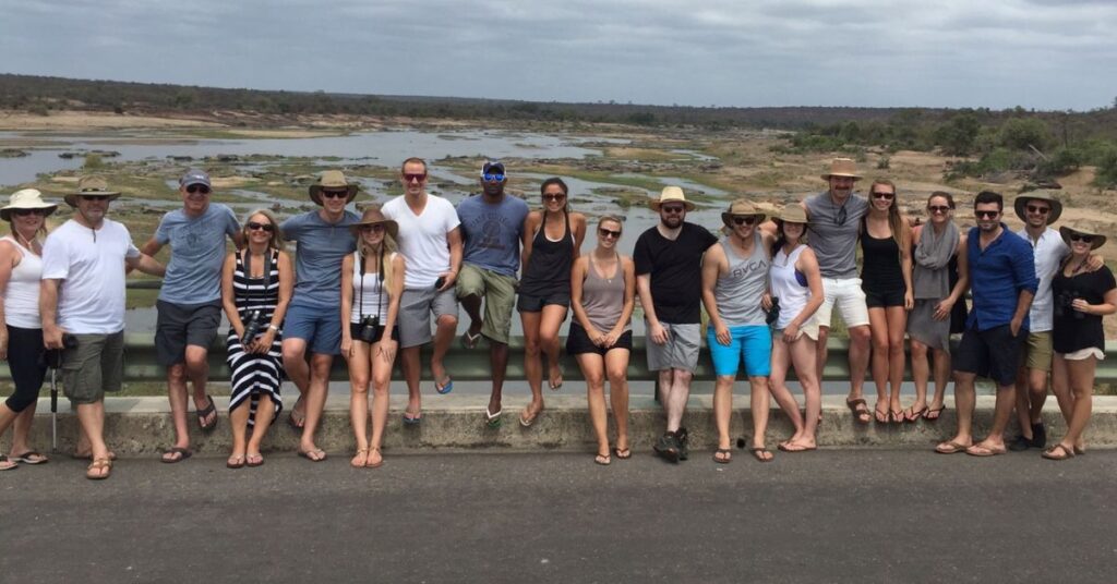 A smiling group of tourists standing together for a photo at a riverside scenic viewpoint during a Kruger National Park safari.