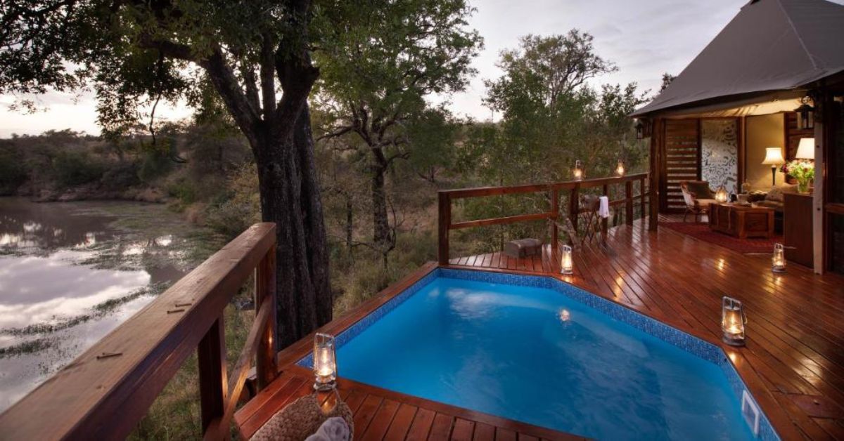 A luxury safari tent opening onto a wooden deck with a built-in plunge pool and lanterns, overlooking a river and lush savanna at Hamiltons Tented Camp.