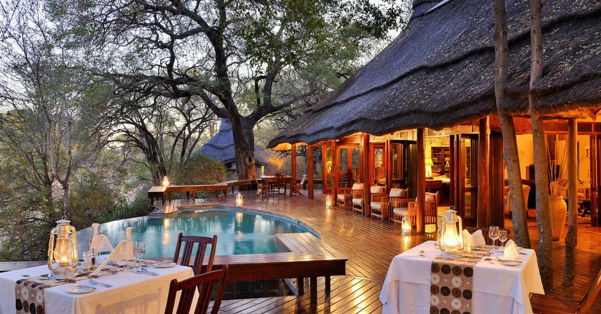 A candlelit dinner setting on a wooden deck with a small swimming pool and a large thatched building under leafy trees at Imbali Safari Lodge.
