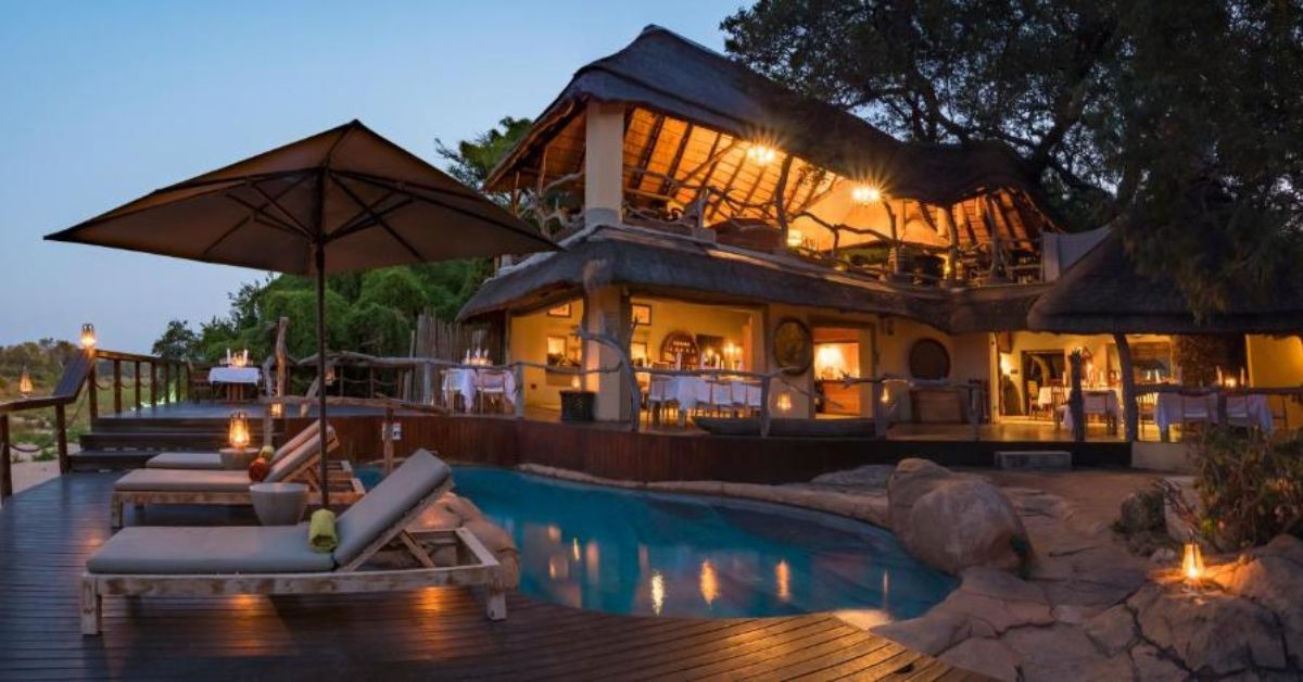 A luxury two-story thatched lodge illuminated at night with a swimming pool, wooden deck, and outdoor dining tables overlooking the bushveld.