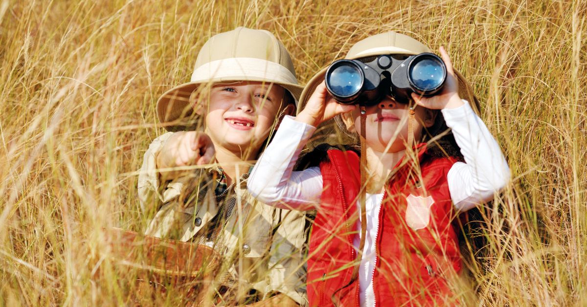 Two young children wearing safari hats exploring the long bushveld grass with binoculars on a kruger safari with kids