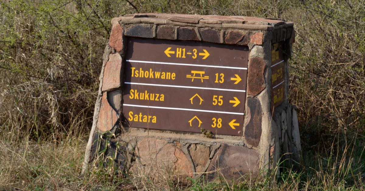 A brown and yellow stone road sign in Kruger National Park showing directions and distances to Tshokwane picnic site, Skukuza rest camp, and Satara rest camp.