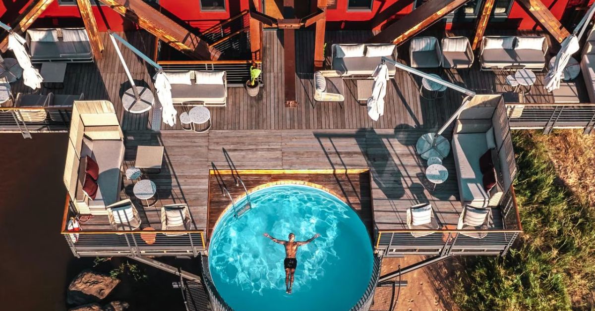 An aerial view of a circular blue swimming pool on a wooden deck suspended over a river, with modern lounge seating and the side of a red train carriage at Kruger Shalati.