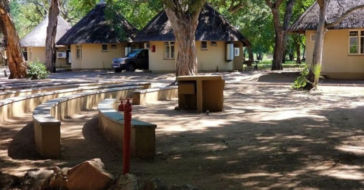 Several cream-colored thatched bungalows under large shady trees at Letaba Rest Camp, with curved stone benches in the foreground.