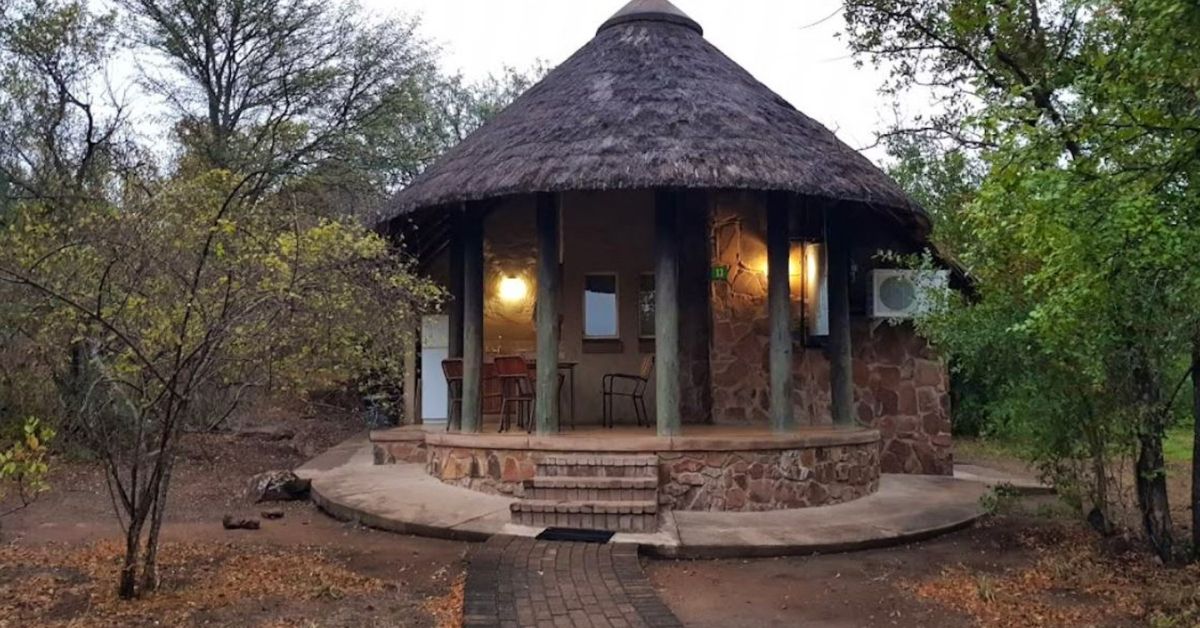 A circular stone-walled chalet with a steep thatched roof and a paved veranda featuring warm outdoor lighting and a dining set at Mopani Rest Camp.