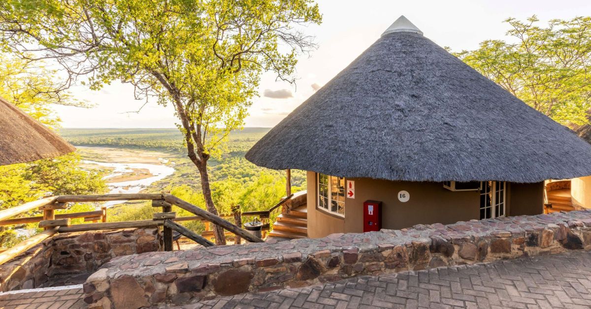 kruger's best camps A traditional round thatched bungalow at Olifants Rest Camp with a stone-walled walkway and a panoramic view of the Olifants River in the background.