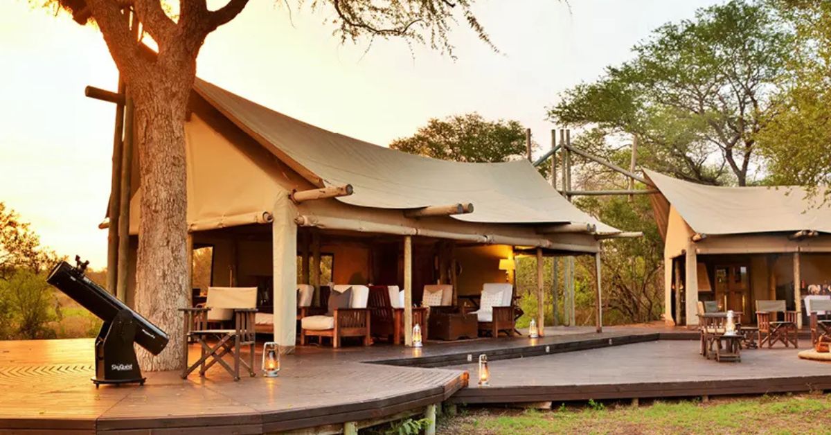 Large safari tents with high canvas roofs opening onto a wide wooden deck with white armchairs, lanterns, and a professional telescope at sunset.