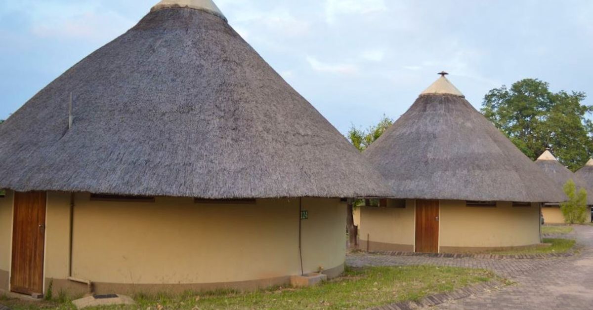 Several traditional circular rondavels with steeply pitched thatched roofs and wooden doors, connected by a paved walkway at Pretoriuskop Rest Camp.