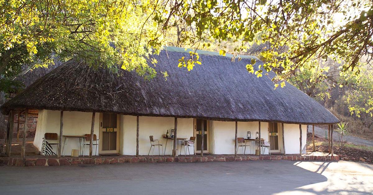 A row of white, traditional thatched-roof bungalows with small outdoor tables and chairs under a deep shaded veranda at Punda Maria Rest Camp.