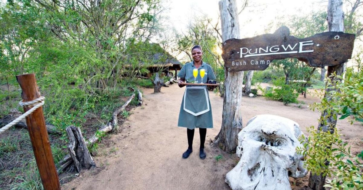 A friendly staff member welcomes visitors at the wooden entrance gate of Pungwe Safari Camp in Manyeleti Game Reserve.