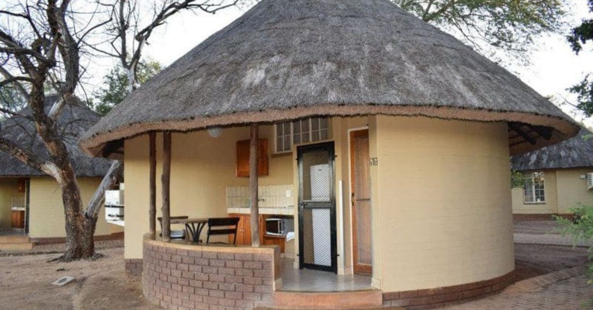 A circular cream-colored bungalow with a thick thatched roof, a private kitchenette, and a small dining table on the porch at Satara Rest Camp.