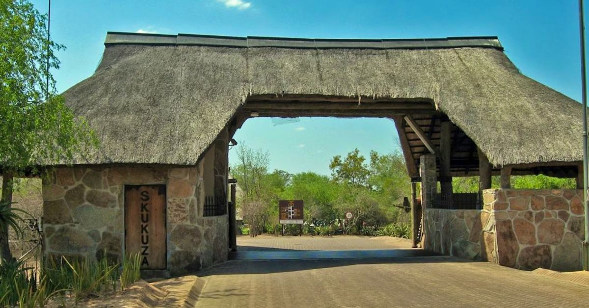 A wide stone entrance gate with a large thatched roof under a clear blue sky at a safari park entrance.