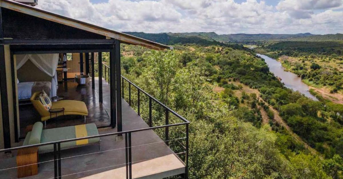 A modern safari suite with an open wall and private balcony featuring sun loungers, offering a panoramic view of a winding river and lush green valley at The Outpost Lodge kruger's best camps